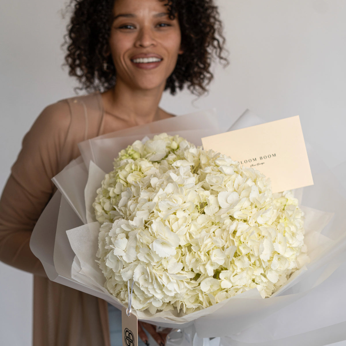 White hydrangea bouquet wrapped in cream paper with Bloom Room card held by smiling woman in beige top