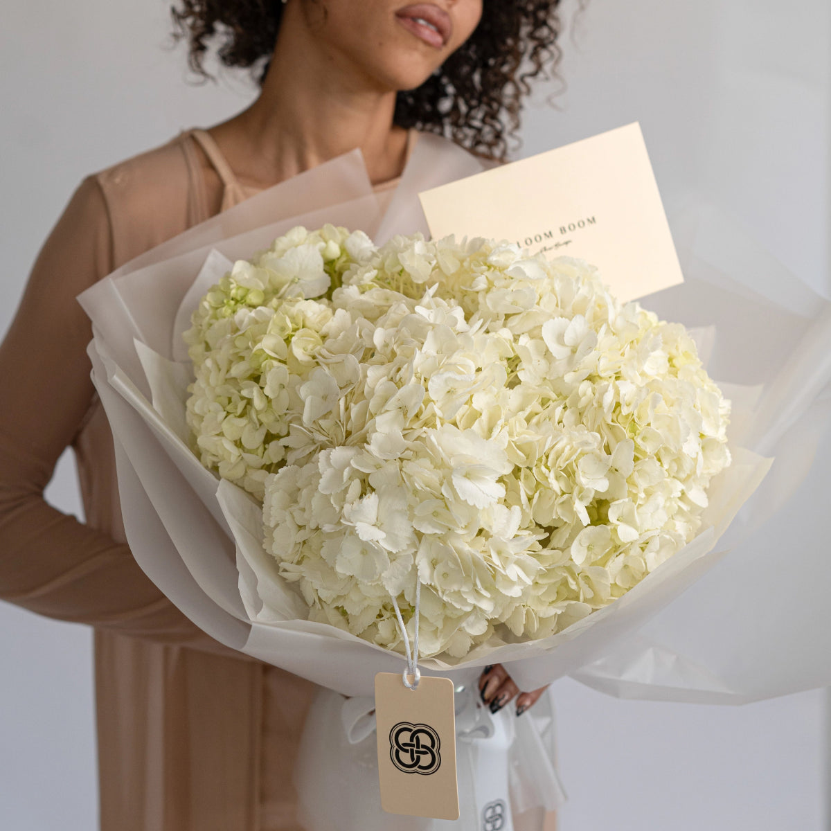 White hydrangea bouquet wrapped in cream paper with gift tag, held by woman in beige sweater