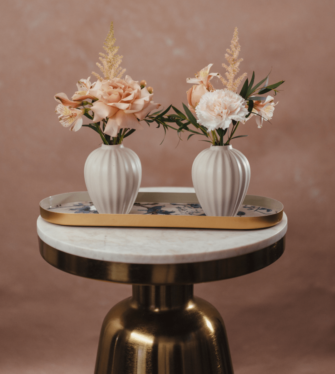 Two white ribbed ceramic vases with pink roses and dried flowers on floral tray atop gold marble side table