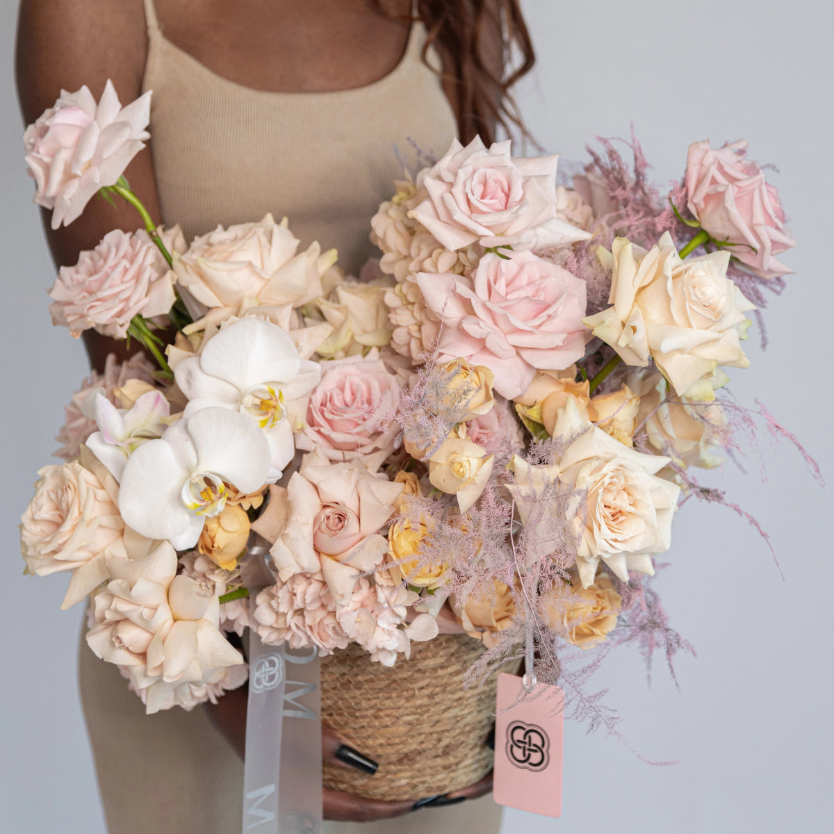 Wicker basket bouquet with pink roses, white orchids, and purple dried grasses with ribbon bow and gift tag